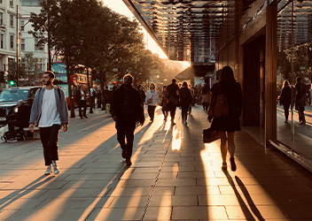 City street scene at sunset with people walking on a sidewalk lined with trees and buildings. The setting sun casts long shadows, and a double-decker bus is visible in the background.